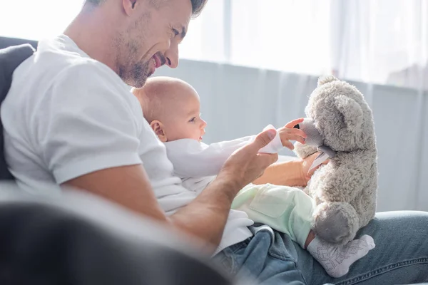 Enfoque selectivo de adorable hija bebé jugando con el oso de peluche y sentado en el sofá con el padre en casa - foto de stock