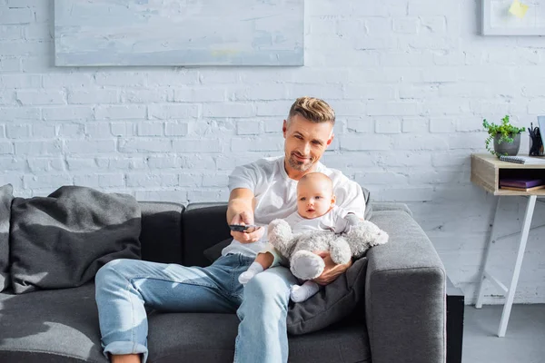 Father sitting on couch with adorable baby daughter, holding remote controller and watching television at home — Stock Photo