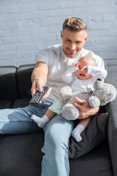 Smiling father sitting on couch with baby daughter, holding remote control and watching television at home — Stock Photo