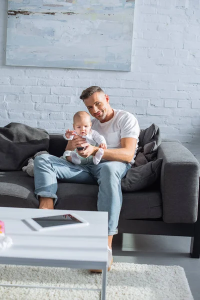 Glücklicher Vater, der mit seiner entzückenden kleinen Tochter auf der Couch sitzt und im Wohnzimmer in die Kamera schaut — Stockfoto