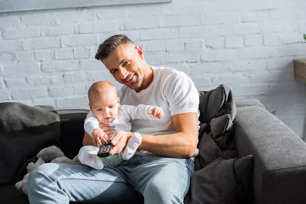 Feliz padre e hija bebé sentado en el sofá, sosteniendo el mando a distancia y viendo la televisión en casa - foto de stock