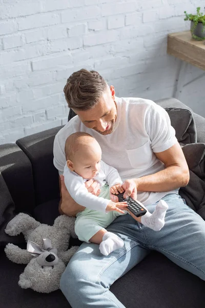Pai e bebê filha sentados no sofá, segurando controlador remoto e assistindo televisão em casa — Fotografia de Stock