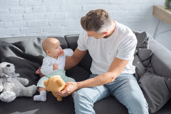 Vater sitzt mit kleiner Tochter auf Couch und spielt zu Hause mit Teddybären — Stockfoto