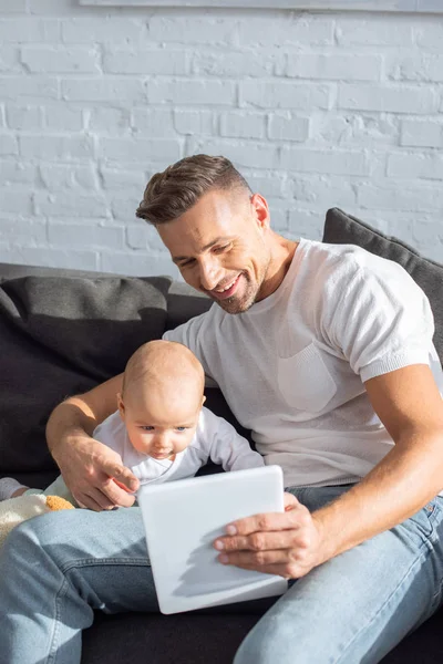 Feliz padre sentado en el sofá con adorable hija bebé y el uso de la tableta digital en casa - foto de stock