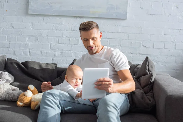 Pai sentado no sofá com a filha do bebê e usando tablet digital na sala de estar — Fotografia de Stock