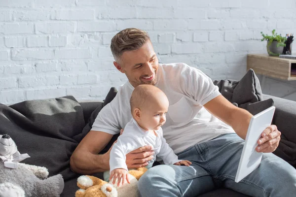 Smiling father sitting on couch with baby daughter and using digital tablet at home — Stock Photo