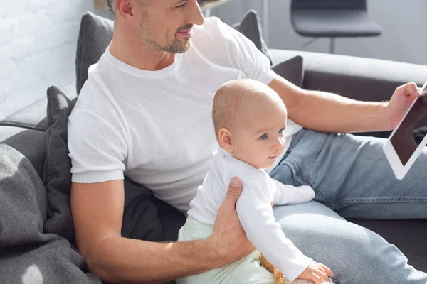 Father sitting on couch with adorable baby daughter and using digital tablet at home — Stock Photo