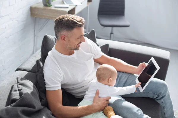 Улыбающийся отец сидит на диване с очаровательной дочкой и использует цифровой планшет с чистым экраном дома — стоковое фото