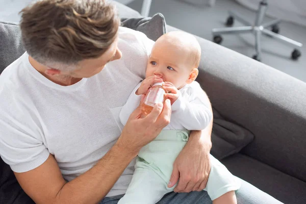 Father sitting on couch and feeding adorable baby daughter from bottle at home — Stock Photo