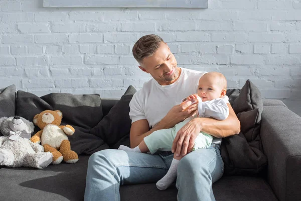 Smiling father sitting on couch and feeding baby daughter in living room — Stock Photo
