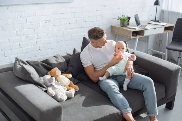 Father sitting on couch and feeding baby daughter from bottle in living room — Stock Photo