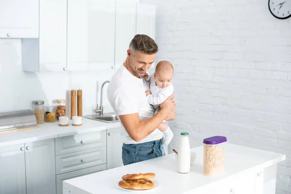 Щасливий батько тримає веселу дитину і дивиться на пляшку з молоком на кухні — стокове фото