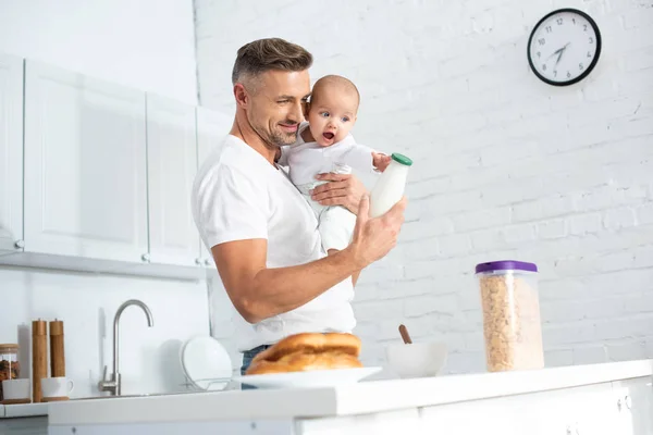 Feliz padre sosteniendo bebé hija y mostrando biberón con leche - foto de stock