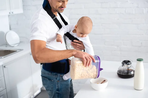 Веселий батько тримає немовляти дочку в дитячому перевізнику і готує сніданок — стокове фото