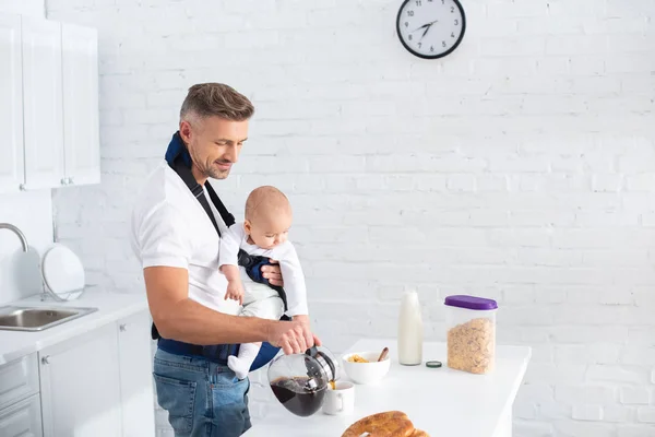 Веселий батько тримає дочку немовляти в дитячому носії і поливає каву в чашку — стокове фото