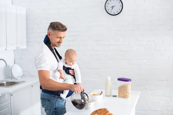 Pai alegre segurando a filha infantil no portador do bebê e derramando café no copo — Fotografia de Stock