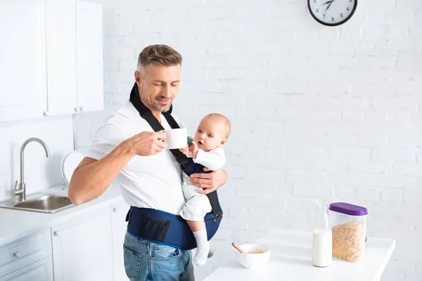 Father holding infant daughter in baby carrier and looking at cup in kitchen — Stock Photo