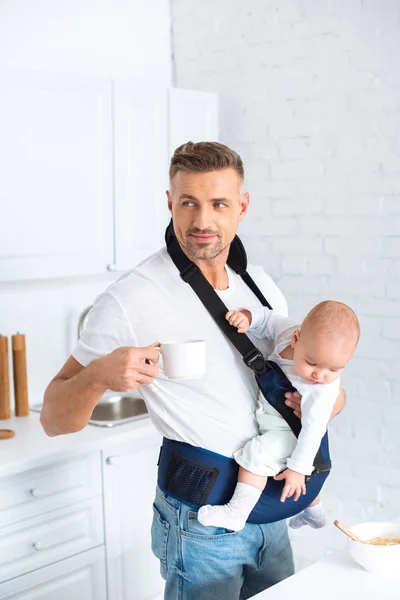 Father holding infant daughter in baby carrier and cup with drink — Stock Photo