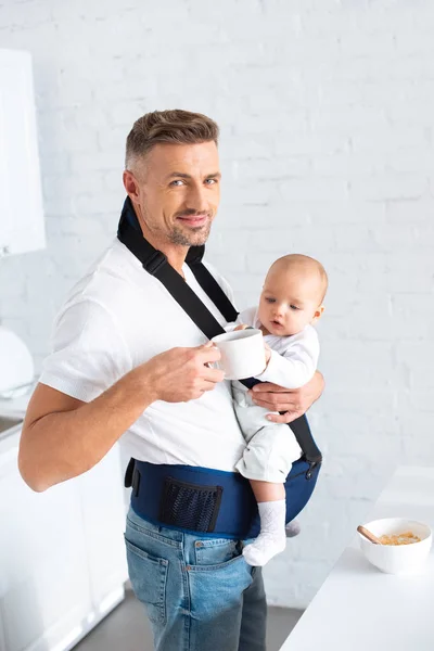 Happy dad holding infant daughter in baby carrier and cup with drink — Stock Photo