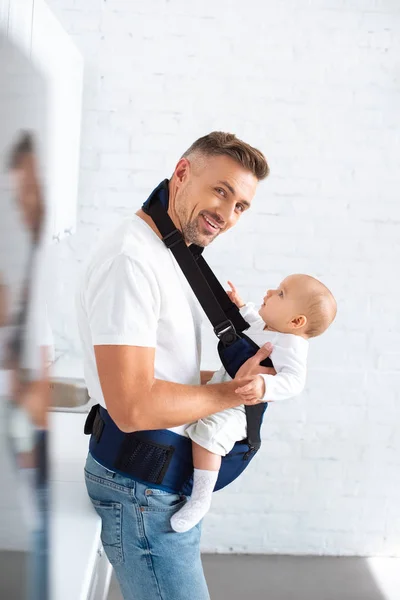Cheerful father holding infant daughter in baby carrier at home — Stock Photo