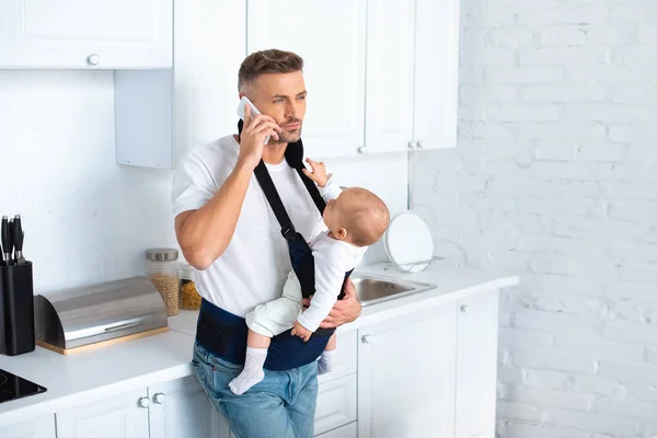 Serious father holding infant daughter in baby carrier and talking on smartphone in kitchen — Stock Photo