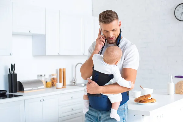 Dad holding infant daughter in baby carrier and talking on smartphone in kitchen — Stock Photo