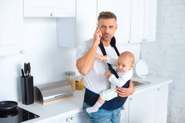 Padre che tiene la figlia neonata nel marsupio e parla sullo smartphone in cucina — Foto stock