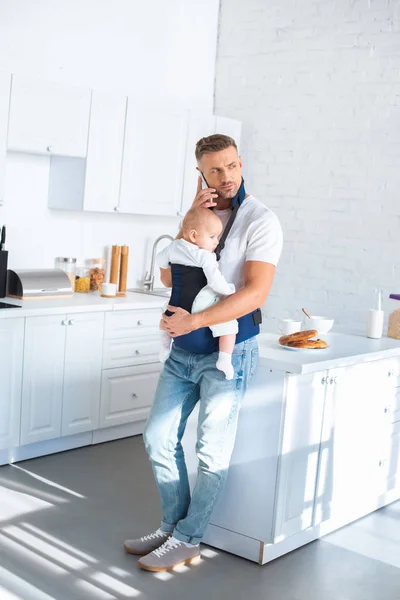 Serious father holding infant daughter in baby carrier and talking on smartphone — Stock Photo