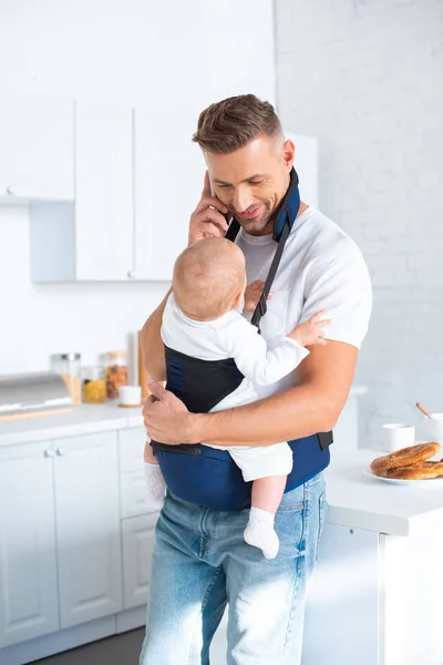 Cheerful father holding infant daughter in baby carrier and talking on smartphone — Stock Photo