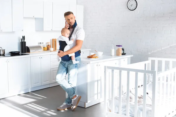 Father holding infant daughter in baby carrier and talking on smartphone — Stock Photo