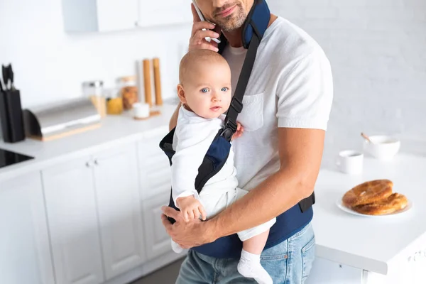 Cropped view of father holding infant daughter in baby carrier and talking on smartphone — Stock Photo