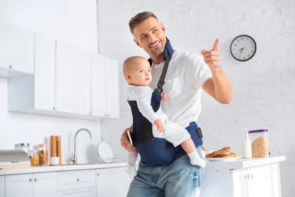 Веселий батько тримає дочку немовляти в дитячому носії і вказує пальцем — стокове фото