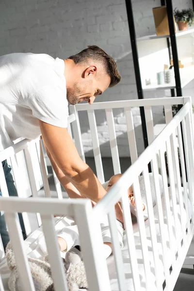 Happy dad putting infant daughter into baby crib at home — Stock Photo