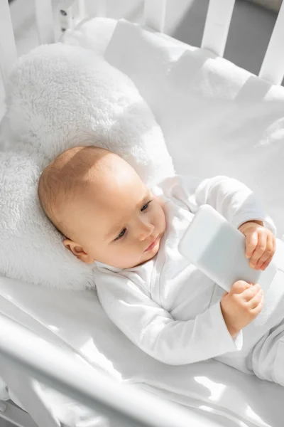 Criança infantil bonito segurando smartphone no berço do bebê branco — Fotografia de Stock