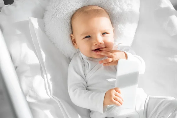 Niño alegre niño sosteniendo smartphone en cuna de bebé blanco - foto de stock