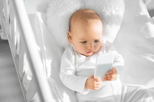 Cute baby holding smartphone in white baby crib — Stock Photo