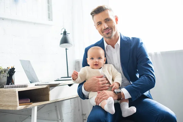 Feliz hombre de negocios sosteniendo hija bebé y sonriendo en la habitación - foto de stock