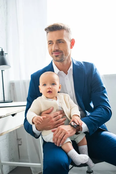 Alegre hombre de negocios sosteniendo hija bebé y sonriendo en la habitación - foto de stock