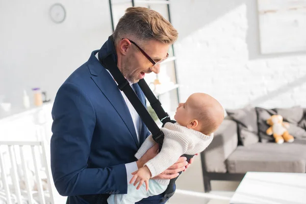 Cheerful businessman looking at infant daughter in baby carrier — Stock Photo
