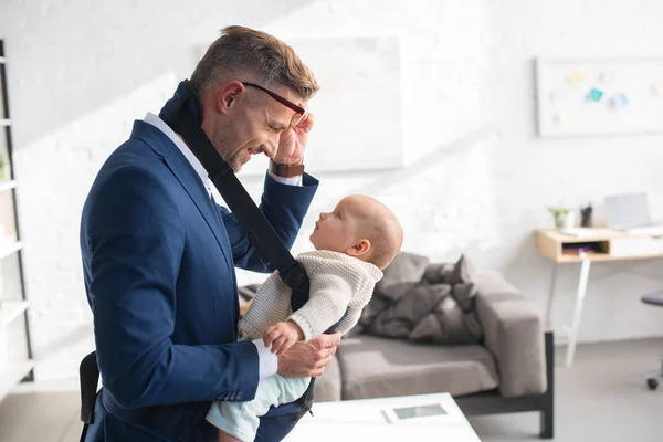Cheerful businessman holding glasses and looking at infant daughter in baby carrier — Stock Photo