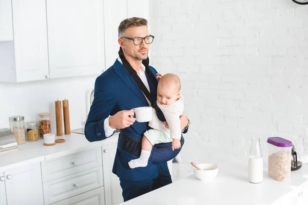 Handsome businessman holding infant daughter in baby carrier and cup with drink — Stock Photo