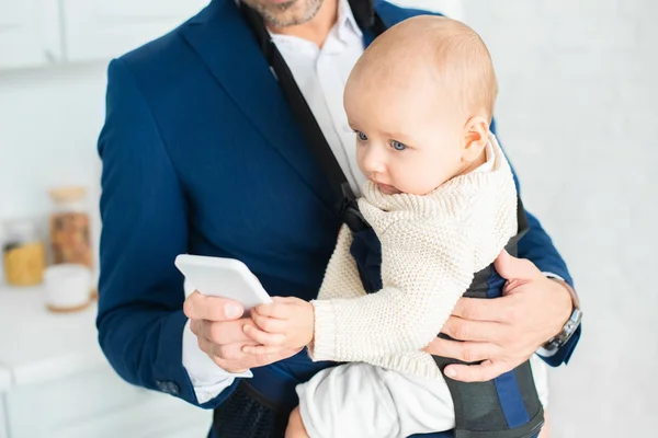 Vista recortada del hombre de negocios sosteniendo teléfono inteligente e hija lactante en portabebés - foto de stock