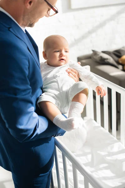 Père en costume tenant la fille de bébé près du lit de bébé dans la chambre — Photo de stock