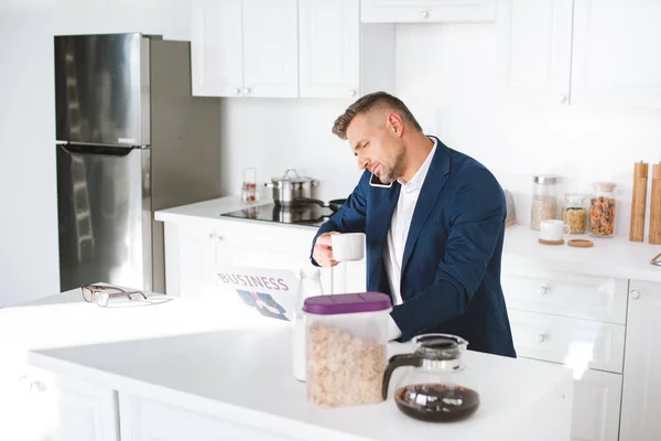 Businessman holding cup with drink while reading business newspaper and talking on smartphone in white kitchen — Stock Photo