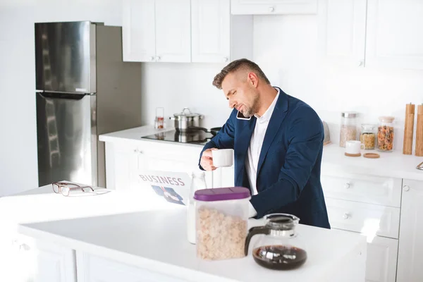 Uomo d'affari in possesso di tazza con bevanda durante la lettura di giornali aziendali e parlare su smartphone in cucina bianca — Foto stock