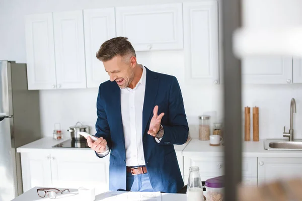 Selective focus of angry businessman yelling while holding smartphone in kitchen — Stock Photo