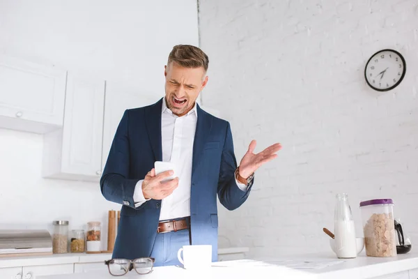 Wütender Geschäftsmann brüllt, während er sein Smartphone in der weißen Küche hält — Stockfoto