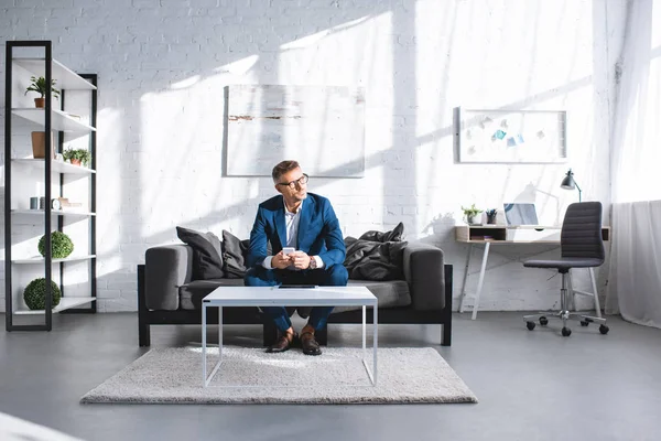 Pensativo hombre de negocios sosteniendo teléfono inteligente y sentado en el sofá en la sala de estar - foto de stock