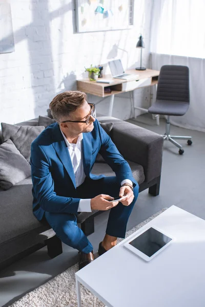 Nachdenklicher Geschäftsmann hält Smartphone in der Hand und sitzt auf Sofa im Wohnzimmer — Stockfoto