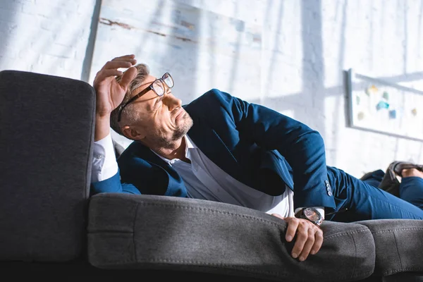 Drunk businessman in glasses lying on sofa with sunshine in living room — Stock Photo